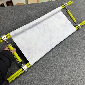 May include: A rectangular embroidery frame with a white fabric stretched across it. The frame has bright yellow bars and silver-colored metal joints. The fabric has a light blue and white pattern. The frame is held by a person.