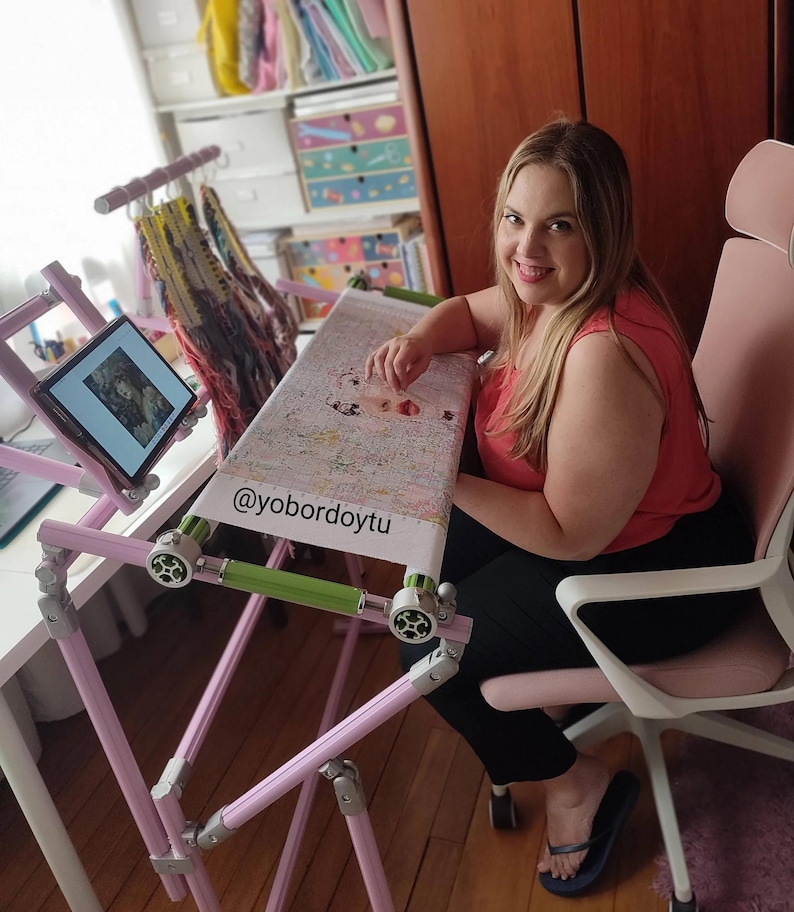 May include: A pink and silver cross-stitch frame with a tablet holder and a cross-stitch project in progress. The frame is adjustable and has a green bar. The text "@yobordoytu" is visible on the project.