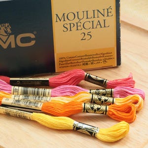 May include: A collection of colorful embroidery floss in shades of pink, orange, and yellow, with a box labeled "MOULINÉ SPÉCIAL 25." The floss is made of 100% cotton and is ideal for various needlework projects.