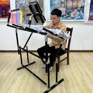 May include: A black and silver adjustable craft stand with a black work surface. The stand has a multi-level design and is being used by a person. A small rack holds colorful thread spools. The stand is on a light wood floor.
