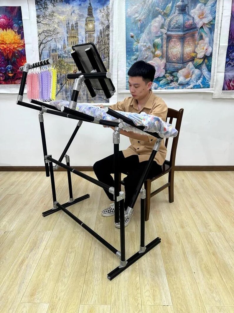 May include: A black and silver adjustable craft stand with a black work surface. The stand has a multi-level design and is being used by a person. A small rack holds colorful thread spools. The stand is on a light wood floor.