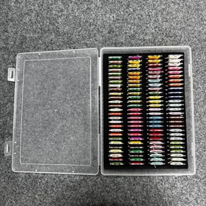 Pode incluir: Um organizador de artesanato de plástico transparente aberto, cheio de bobinas de linha coloridas. As bobinas estão dispostas em fileiras em uma inserção de espuma preta. Em uma superfície cinza.