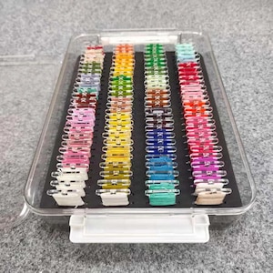 Pode incluir: Um organizador de plástico transparente cheio de carretéis de linha colorida. A linha é disposta em fileiras verticais de várias cores, incluindo rosa, amarelo, verde, azul e vermelho. O organizador tem um interior preto e uma trava branca.