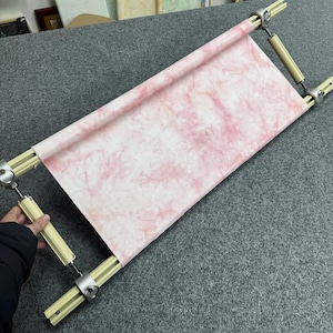 May include: A wooden embroidery hoop with a pink and white fabric stretched across it. The fabric has a tie-dye pattern. The frame is made of light-colored wood and metal hardware. The hoop is designed to hold fabric taut for needlework.