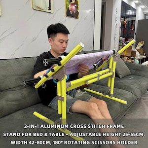 May include: A yellow aluminum cross stitch frame with a purple fabric stretched across it. The frame is adjustable, with a height of 9.8-21.6 inches and a width of 16.5-31.5 inches. The frame includes a 180° rotating scissors holder. The text on the image reads: 2-IN-1 ALUMINUM CROSS STITCH FRAME STAND FOR BED & TABLE - ADJUSTABLE HEIGHT 25-55CM, WIDTH 42-80CM, 180° ROTATING SCISSORS HOLDER.
