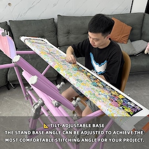 May include: A light purple, tilt-adjustable embroidery stand with a cross-stitch project in progress. The stand's base angle can be adjusted for comfortable stitching. The text on the image reads: "TILT-ADJUSTABLE BASE THE STAND BASE ANGLE CAN BE ADJUSTED TO ACHIEVE THE MOST COMFORTABLE STITCHING ANGLE FOR YOUR PROJECT."