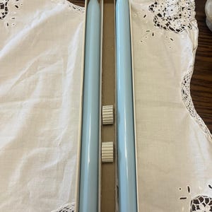 May include: Two light blue candles in a white box. The candles are long and thin, and they are packaged together in a white box with a clear plastic window.