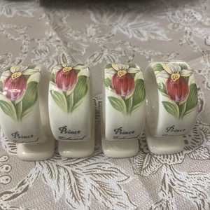 May include: Four ceramic napkin rings with a floral design. Each ring features a red and yellow flower with green leaves. The word "Prince Edward" is printed on each ring in black script. The rings are on a patterned tablecloth.