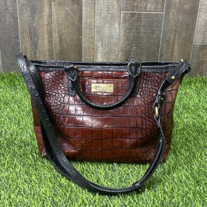 May include: A brown handbag with a textured crocodile-embossed pattern, featuring black leather trim, two top handles, and an adjustable shoulder strap. A rectangular gold-tone metal plate with engraved text is centered on the front panel.