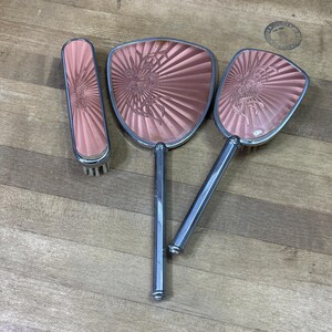 Puede incluir: Un juego de tocador que consta de un cepillo rectangular, un espejo de mano y un cepillo para el cabello, todos con un acabado metálico en oro rosa con un patrón de rayos y un diseño floral en relieve. Cada pieza tiene un mango plateado.