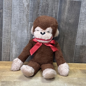 May include: A plush monkey toy with dark brown fur, a light beige face, hands, and feet. The monkey features a stitched black nose and mouth, and a bright red satin ribbon tied in a bow around its neck. The toy is seated in an upright position.
