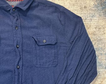 May include: A navy blue button-down shirt with a chest pocket. The shirt has a collared neckline and long sleeves. The buttons are dark, and the fabric appears to be a soft, woven material. The shirt is a casual style.