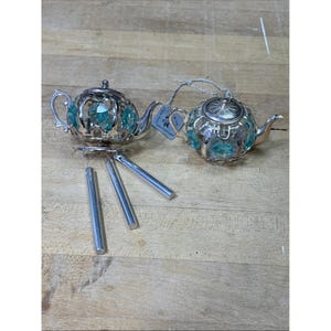 Può includere: Due ornamenti a forma di teiera in metallo color argento con cornici traforate e cristalli sfaccettati color turchese incastonati. Una teiera include tre campanelli cilindrici argentati pendenti dalla base, l'altra ha un occhiello per appenderla.