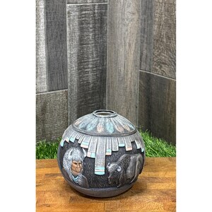 May include: A spherical ceramic vase in a dark gray finish featuring a relief design of a person wearing a feathered headdress and a buffalo. The upper portion is decorated with a band of carved feathers accented with light blue and tan paint.