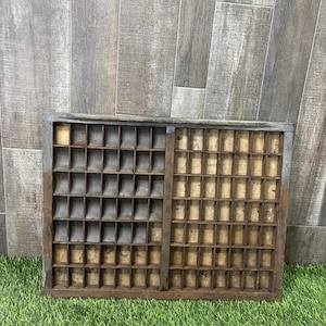 May include: A rectangular wooden printer's tray featuring a grid of numerous small, open compartments. The left side contains curved dividers, while the right side has straight dividers. The wood has a dark, weathered finish with visible grain.