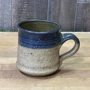 May include: A ceramic mug featuring a two-tone glaze design. The upper portion is a deep, glossy navy blue, while the lower body and handle exhibit a textured, speckled beige and tan finish. The mug has a wide, rounded base and a sturdy handle.