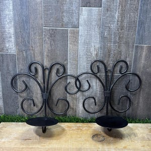 Puede incluir: Un par de apliques de pared de metal negro con diseños de volutas simétricas. Cada aplique tiene una base plana circular para sostener una vela y una placa posterior vertical con detalles de alambre curvado en un patrón decorativo.