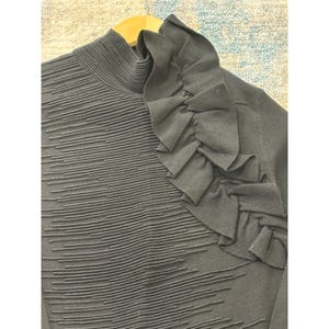 May include: A black sweater with a high, ribbed collar and a textured, horizontal striped pattern on the front. The shoulder features a large, ruffled design. The sweater is hanging on a wooden hanger.