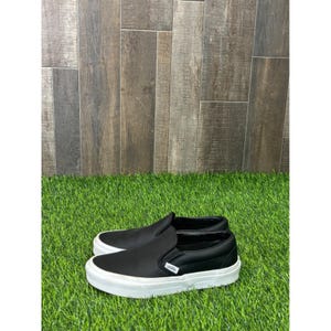 Op de afbeelding: Zwarte slip-on sneakers met witte zolen. De sneakers hebben een geperforeerd ontwerp op de bovenkant en het Vans-logo aan de zijkant. De schoenen worden getoond op een bed van kunstgras tegen een achtergrond van houtnerfpanelen.