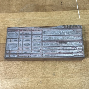 May include: A rectangular metal printing plate featuring a grid layout for financial record keeping. The surface contains embossed text including labels like DATE, PAYMENT, and INTEREST, with various blank rectangular fields for data entry.