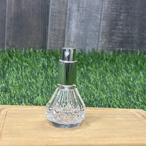 Puede incluir: Un frasco de perfume de vidrio transparente y facetado con un pulverizador metálico plateado y verde. El frasco tiene un patrón de diamantes y se encuentra sobre una superficie de madera. El fondo presenta hierba verde y una pared de madera.
