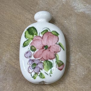 May include: A white ceramic pendant with a flat, oval shape featuring a hand-painted design of a large pink flower, smaller purple blossoms, and green leaves. The piece has a small, rounded protrusion at the top and a smooth, glossy finish.