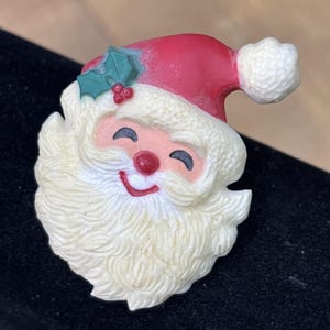 May include: A Santa Claus brooch featuring a red hat with a white pom-pom and a green holly leaf with red berries. The face has a textured white beard, a pink complexion, a red nose, and a smiling expression with closed eyes and a red mouth.