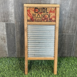 Op de afbeelding: Een vintage houten wasbord met een metalen schrobvlak. Het bovenste gedeelte toont de woorden "DUBL HANDI" in rode letters, samen met extra tekst. Het wasbord is gemaakt van lichtgekleurd hout en staat op een grasachtig oppervlak.