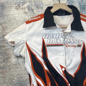 May include: White short-sleeved button-up shirt with black collar and orange and black flame design. The shirt features the Harley-Davidson Racing logo and the Harley-Davidson name printed on the front and shoulder.