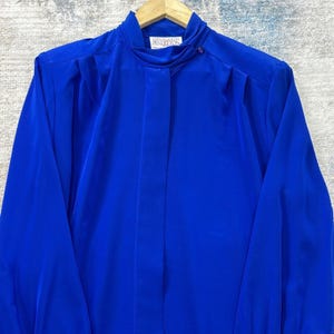 May include: A vibrant blue blouse with a high collar and long sleeves. The blouse features a button closure at the neck and pleats at the shoulders. The brand name "Pendleton" is visible on the label. The fabric has a smooth, flowing texture.