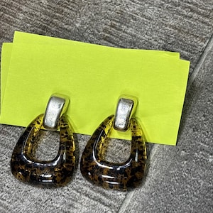May include: A pair of earrings with a silver top and a brown and black spotted, teardrop-shaped bottom. The earrings are displayed on a bright yellow sticky note, set against a gray surface.