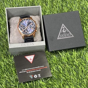 May include: A Guess watch with a rose gold case, black leather strap, and a blue dial with Roman numerals. The watch is in a grey box with the Guess logo, with a black box and a card with text.