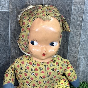 May include: Vintage doll with a painted face, blue eyes, and an open mouth. The doll wears a yellow dress and bonnet patterned with red hearts and keys. The doll's arms and legs are a blue fabric.