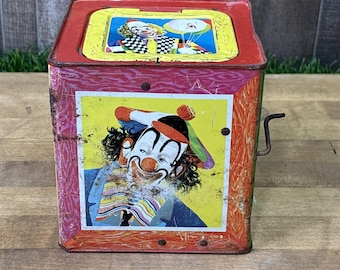 May include: A vintage metal jack-in-the-box toy featuring a red frame with a yellow top panel and a side crank. The front panel displays a colorful illustration of a clown with a red nose, wearing a multi-colored hat and a striped collar.