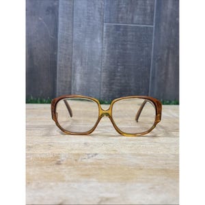 May include: A pair of eyeglasses with a translucent amber-colored plastic frame. The lenses are clear and feature a visible horizontal line indicating bifocal segments. The frame has a rounded rectangular shape with thick rims and thin temples.