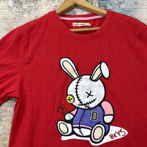 Può includere: T-shirt rossa con un disegno di coniglio dei cartoni animati. Il coniglio ha un aspetto cucito, tiene in mano una rosa e indossa una giacca viola con una "B". Il testo "BKYS" è sotto il coniglio.