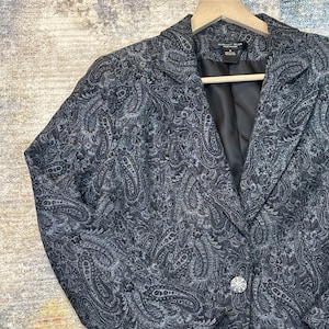 Puede incluir: Un blazer gris oscuro con un estampado de cachemira. El blazer tiene solapas con muescas, dos botones y un forro negro. El blazer está colgado en una percha de madera.