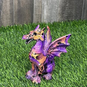 May include: A detailed, colorful dragon figurine with a purple body, orange accents, and large wings. The dragon is posed on a bed of green grass, with a wooden fence in the background. The dragon's wings are a mix of purple and orange.
