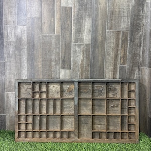 May include: A rectangular wooden printer's tray featuring a grid of numerous small, open compartments of varying sizes. The wood has a weathered, dark brown finish with visible signs of wear, including patches of peeling surface texture.