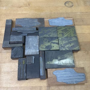 May include: A collection of rectangular metal and wood letterpress printing blocks of various sizes. The blocks feature engraved imagery, including architectural structures, human portraits, and text, with some blocks showing metallic surfaces.