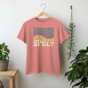 May include: A pink t-shirt with a retro-style graphic design featuring the words "Einkorn", "Khorasan", "Durum", and "Spelt" in a colourful, wavy font.