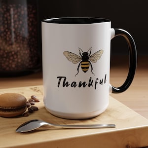 May include: White ceramic mug with a black interior and handle. The mug features a detailed illustration of a bee with the word "Thankful" in a handwritten font. A spoon, a macaron, and coffee beans are on a wooden surface.