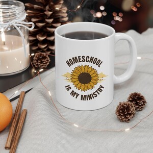 Könnte beinhalten: Weiße Keramik-Kaffeetasse mit dem Text "Homeschool is my Ministry" und einer Sonnenblumen-Grafik. Die Tasse ist mit einer dunklen Flüssigkeit gefüllt und hat einen weißen Henkel. Die Tasse steht auf einer hellgrauen Oberfläche.
