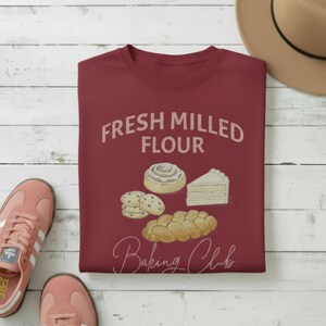May include: A maroon crew neck sweatshirt with the words "FRESH MILLED FLOUR" and "Baking Club" printed on it. The graphic includes illustrations of baked goods like a cinnamon roll, cookies, a slice of cake, and a loaf of bread. Pink trainers and a tan hat are also visible.
