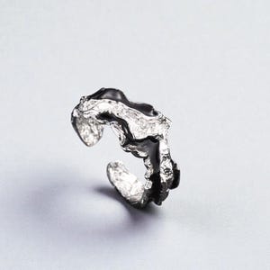 Op de afbeelding: Een zilverkleurige ring met een uniek, organisch ontwerp. De ring heeft een getextureerde, gesmolten-achtige uitstraling met een donkere, glanzende inleg. Het open ontwerp maakt een verstelbare pasvorm mogelijk.