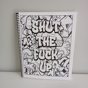May include: A spiral-bound notebook with a black and white graffiti-style design. The text "SHUT THE FUCK UP" is prominently displayed in bold, stylized lettering. The background is filled with abstract shapes and patterns.