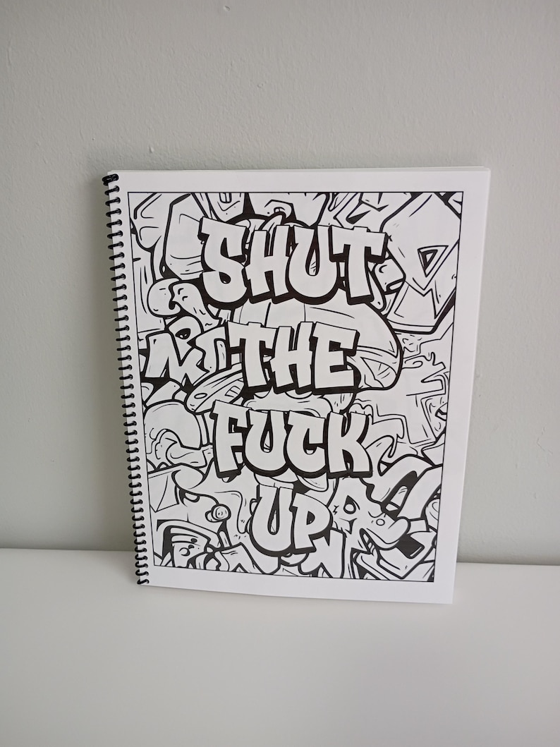 May include: A spiral-bound notebook with a black and white graffiti-style design. The text "SHUT THE FUCK UP" is prominently displayed in bold, stylized lettering. The background is filled with abstract shapes and patterns.