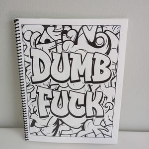 May include: A spiral-bound notebook with a black and white graffiti-style design. The words "DUMB FUCK" are prominently displayed in bold, bubble letters against a backdrop of abstract shapes and lines.