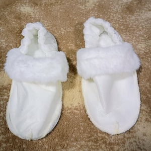 May include: A pair of white slippers with a fluffy, faux fur trim around the opening. The slippers have a soft, textured fabric and a thong-style design. The background is a brown, textured surface.
