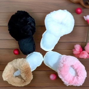 May include: Four fluffy slippers in black, white, tan, and pink are arranged on a wooden surface. Each slipper has a soft, faux fur upper and a white sole. Decorative red spheres and pink flowers are also visible.